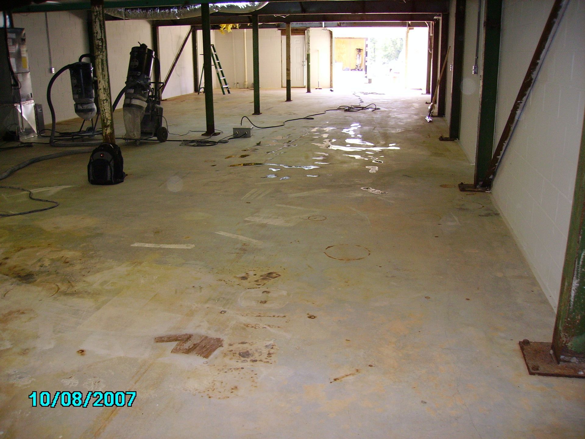 A flooded concrete basement with support beams and an open doorway. Black pump, tools, and stains cover the floor.