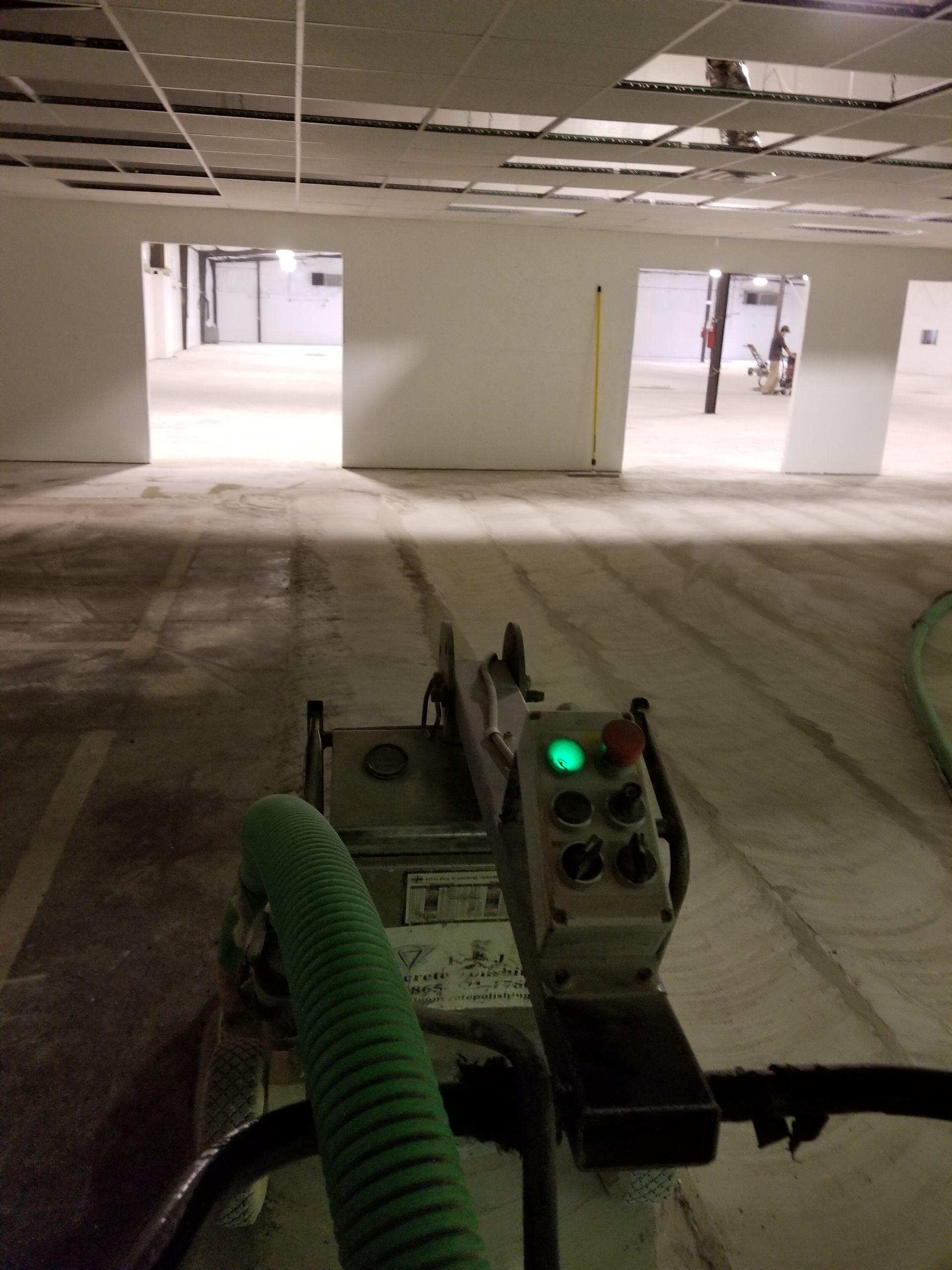 A person operating a concrete grinder in an unfinished building. The grinder has a green hose, a control panel, and grinds the concrete floor.