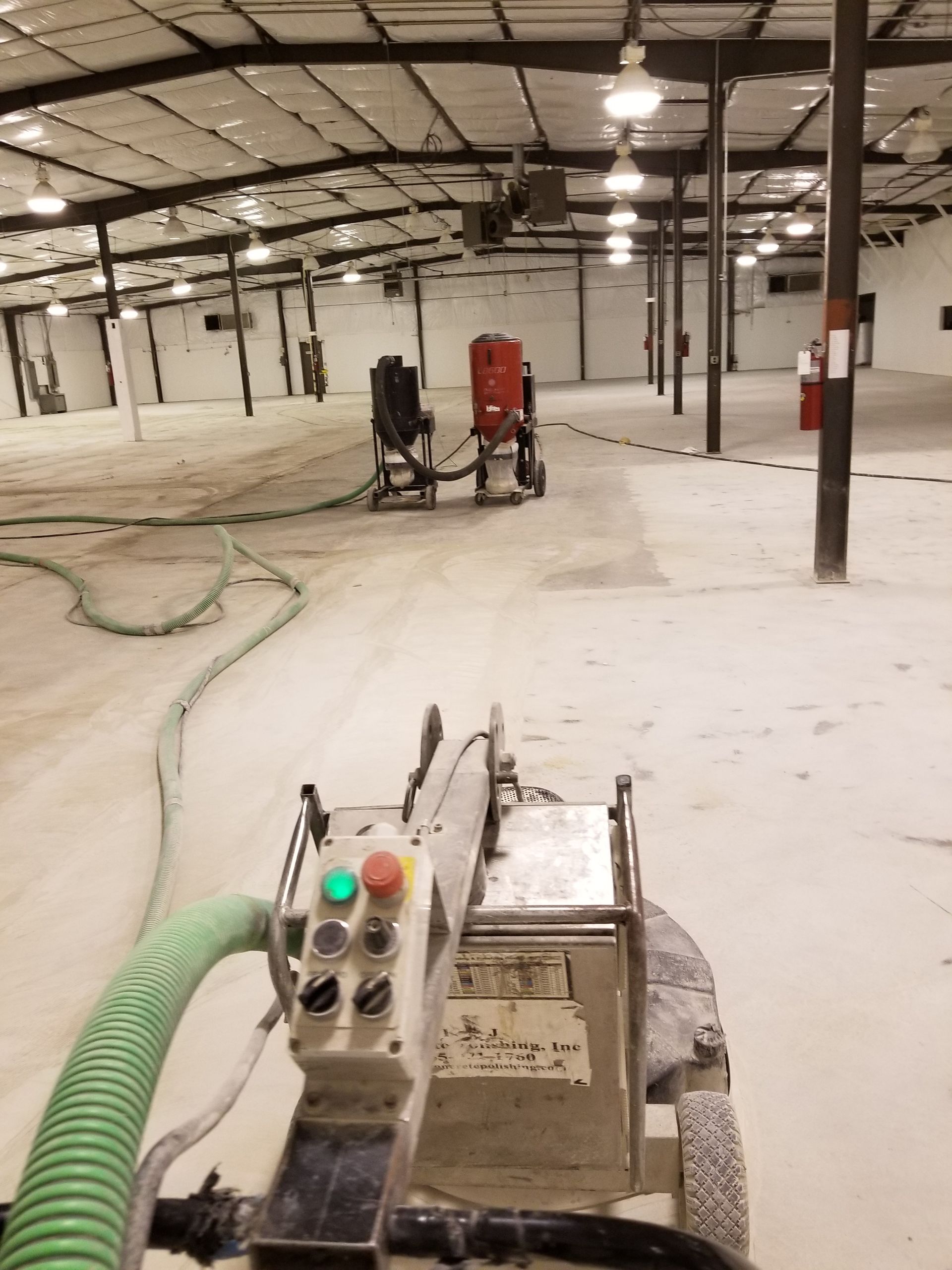 An industrial floor being prepared with a cutting machine and sandblasting equipment. A long warehouse space with white floors and support columns.