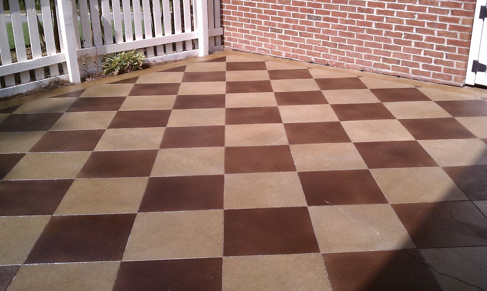 Checkerboard patio made of brown and tan squares, next to a white picket fence and brick wall.