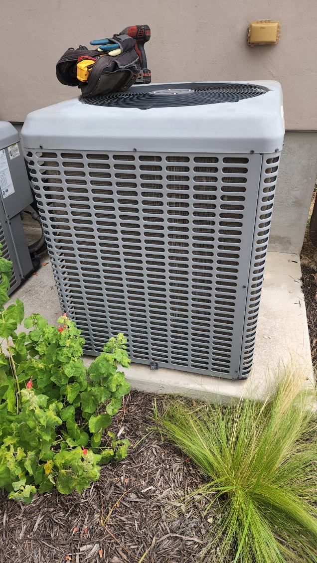 A large air conditioner is sitting on the side of a building next to a bush.