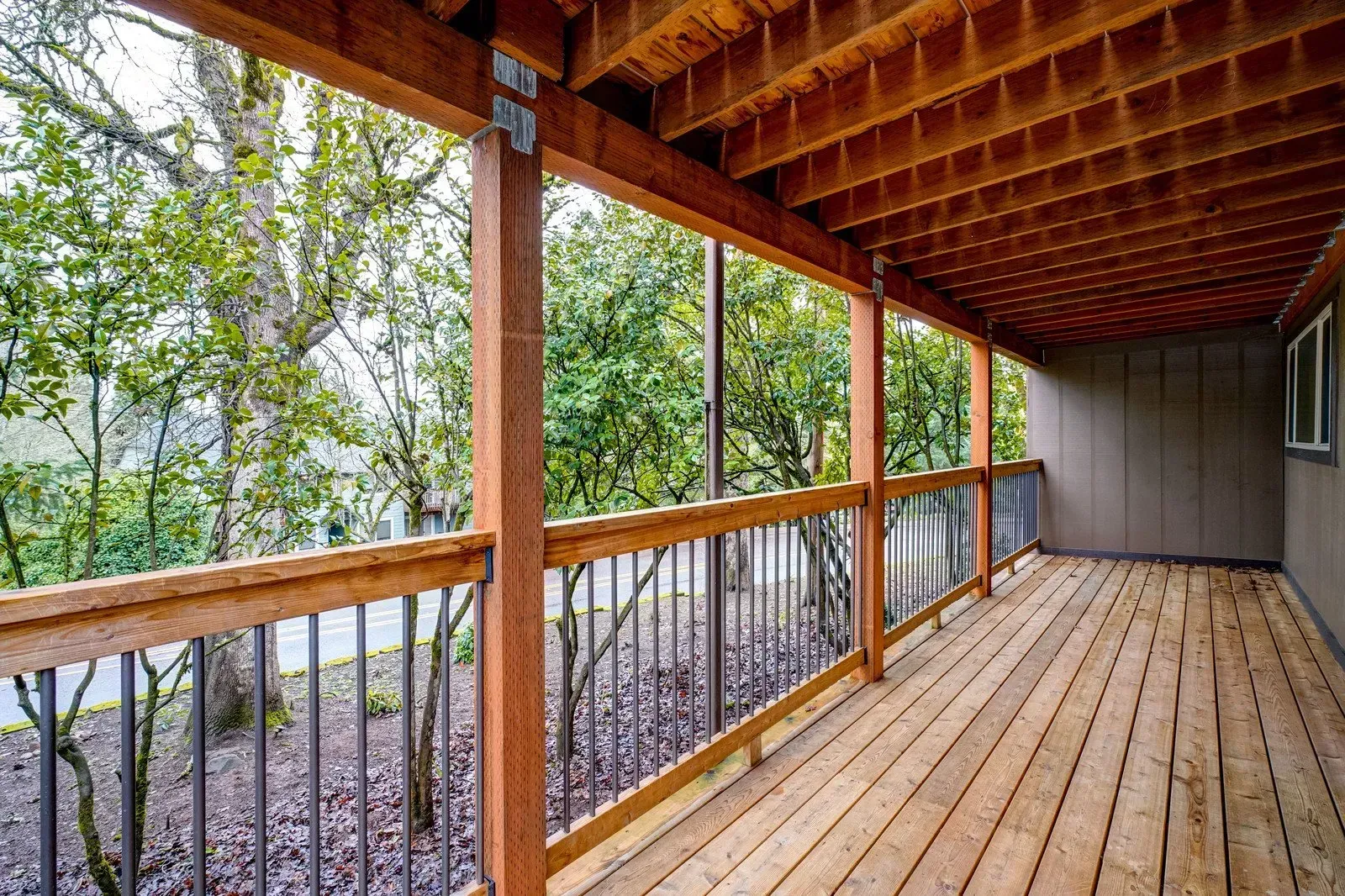Wooden deck with railing, overlooking trees and a path, under a covered roof.