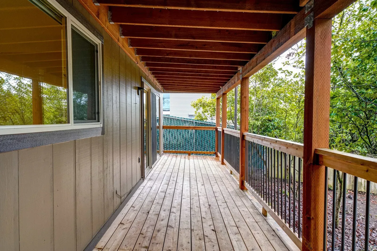 Covered wooden porch with wooden beams, railing, and decking. Trees visible in the background.