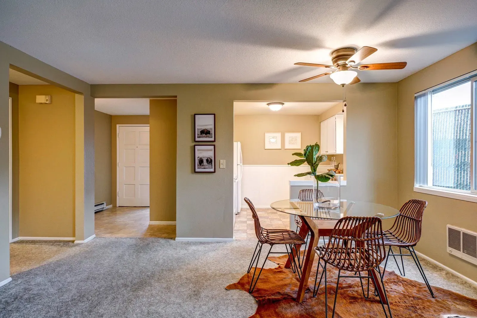 Dining room with table, chairs, and rug, open to a kitchen. Beige walls and gray carpet.