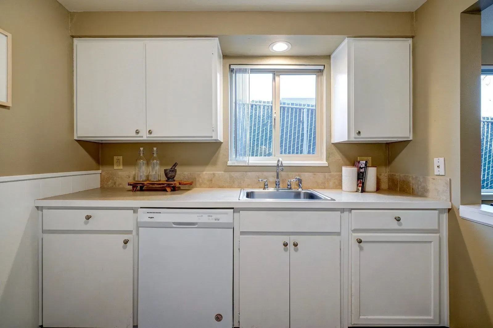 White kitchen cabinets above and below a sink, with a window and dishwasher.