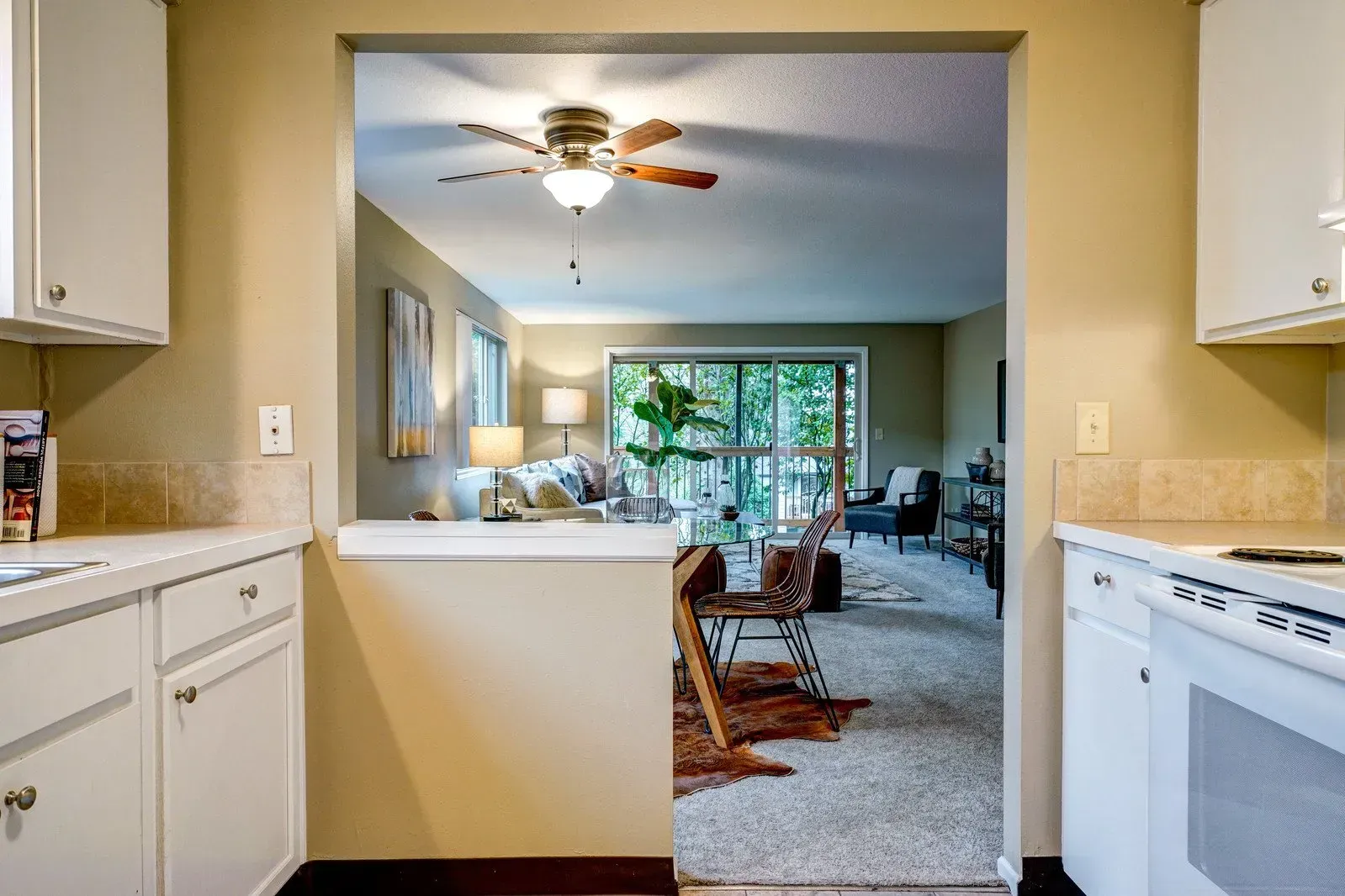 Kitchen doorway opening to a living room with white cabinets, beige walls, and a ceiling fan.