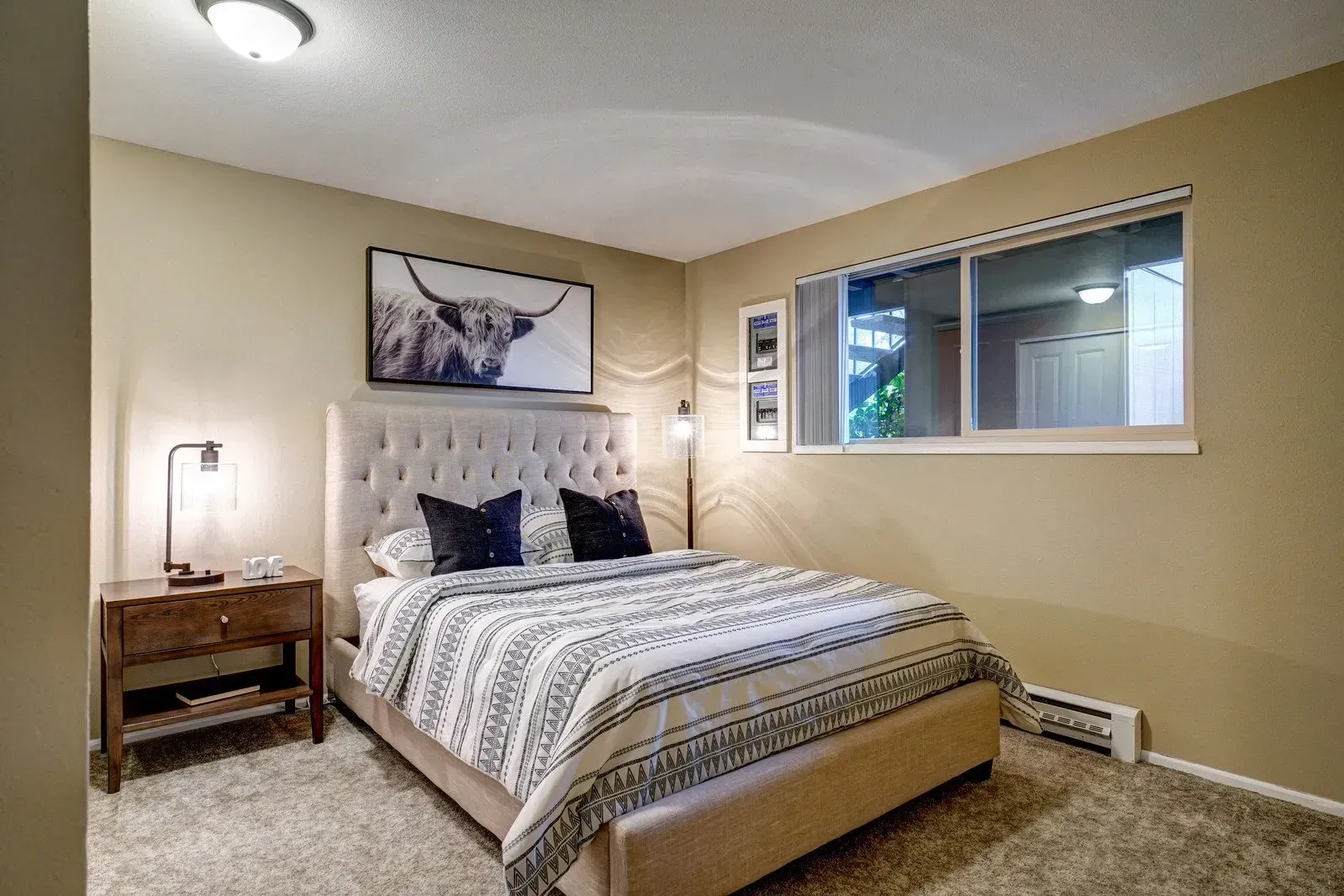Bedroom with bed, nightstand, artwork, and window. Beige walls and carpet.