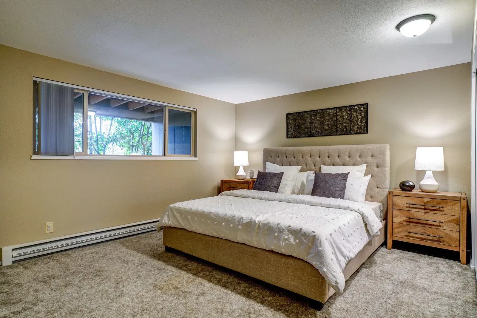 Bedroom with a beige bed, two bedside tables with lamps, a window, and beige walls and carpet.