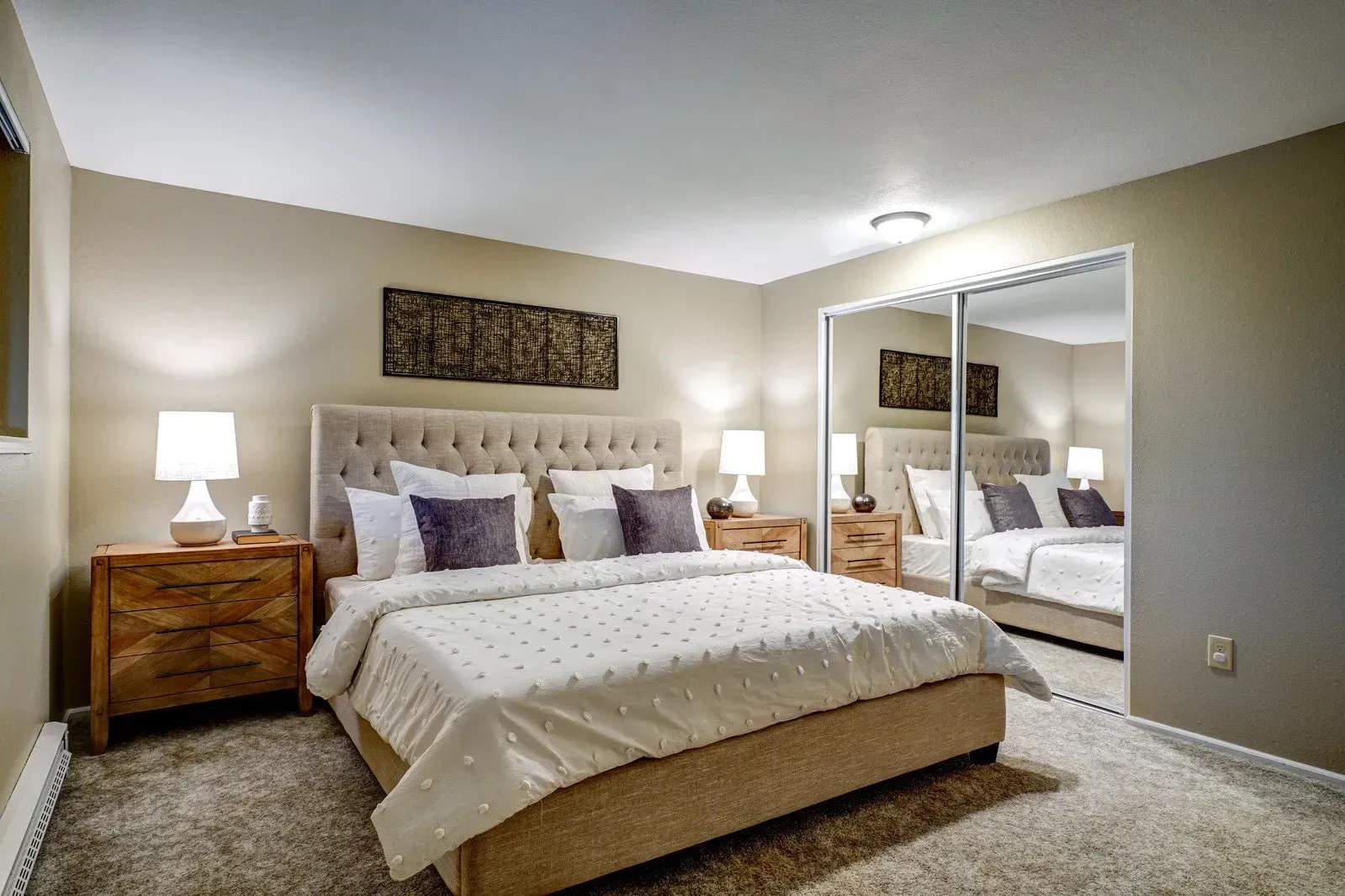 Bedroom with a bed, nightstands, lamps, and a mirrored closet against beige walls, and plush carpet.