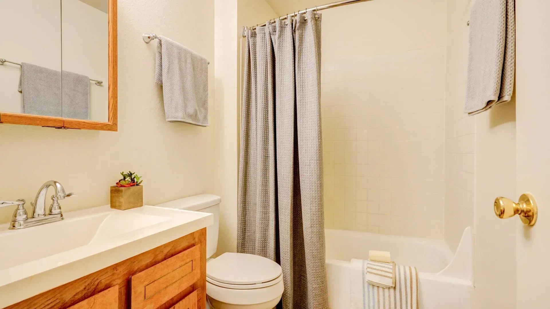 Bathroom with a sink, toilet, tub, and shower curtain. Light beige walls, wooden cabinets, and a framed mirror.