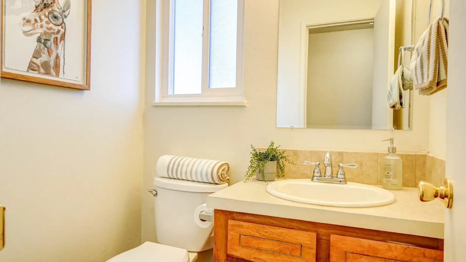 Small bathroom with a toilet, sink, mirror, small window, and giraffe artwork. Light beige walls.