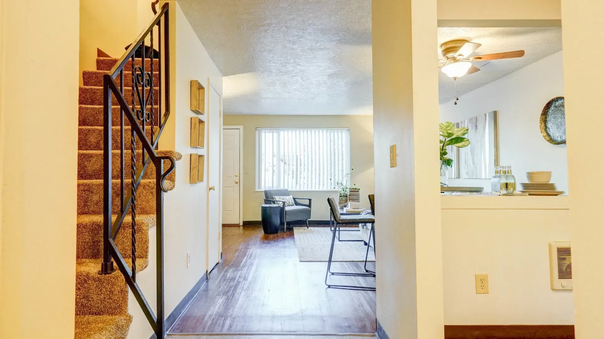 Staircase with brown carpet and black railing leads to a hallway. Living area with table and chairs visible at the end.