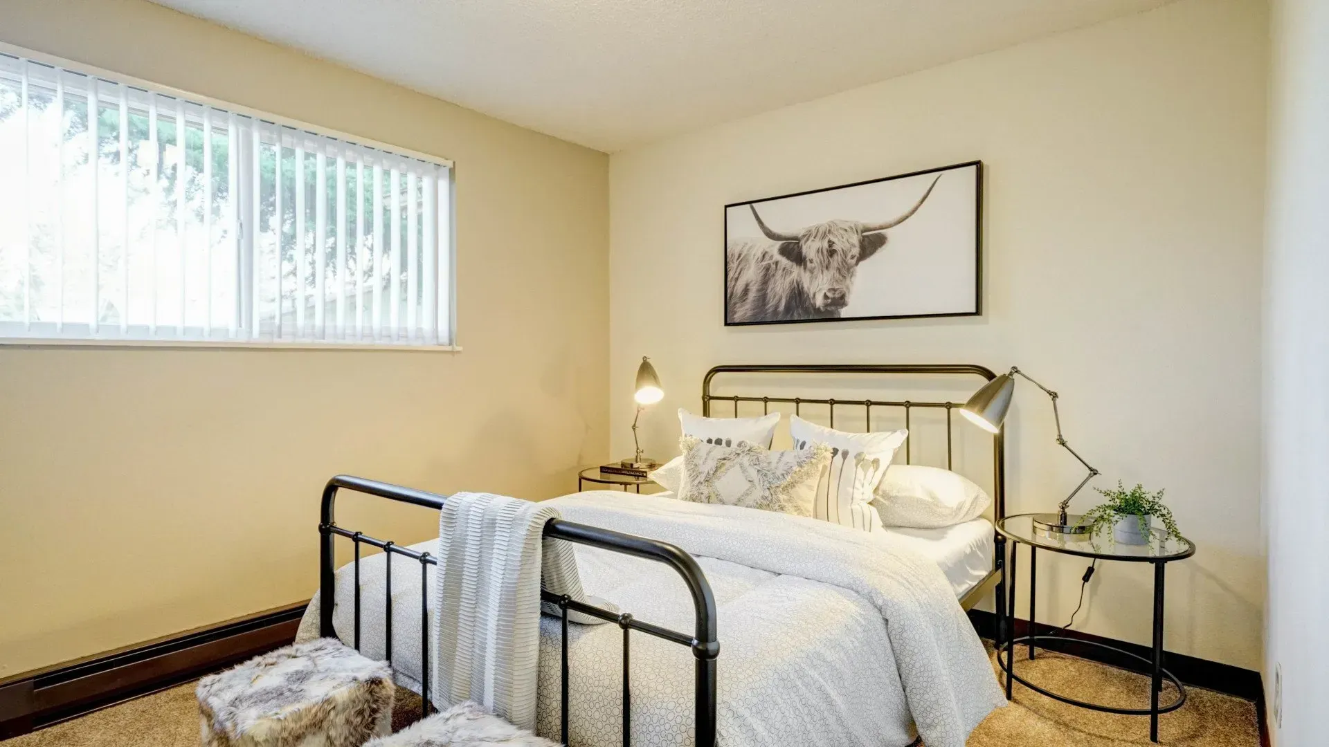 Bedroom with metal bed frame, framed cow art, side table, and window with blinds.