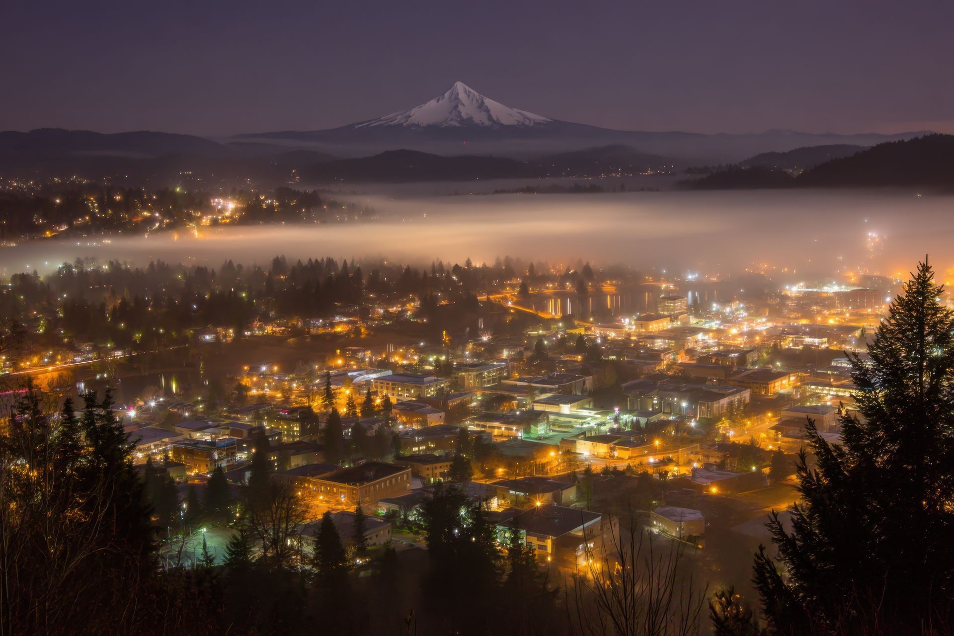 City lights below fog at dusk, Mount Hood in the background.