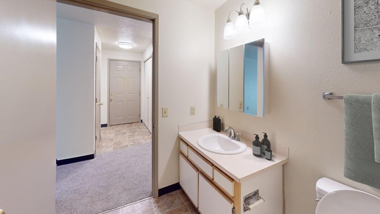Bathroom with a sink, mirror, and toilet. A doorway leads to a hallway with a closed door.