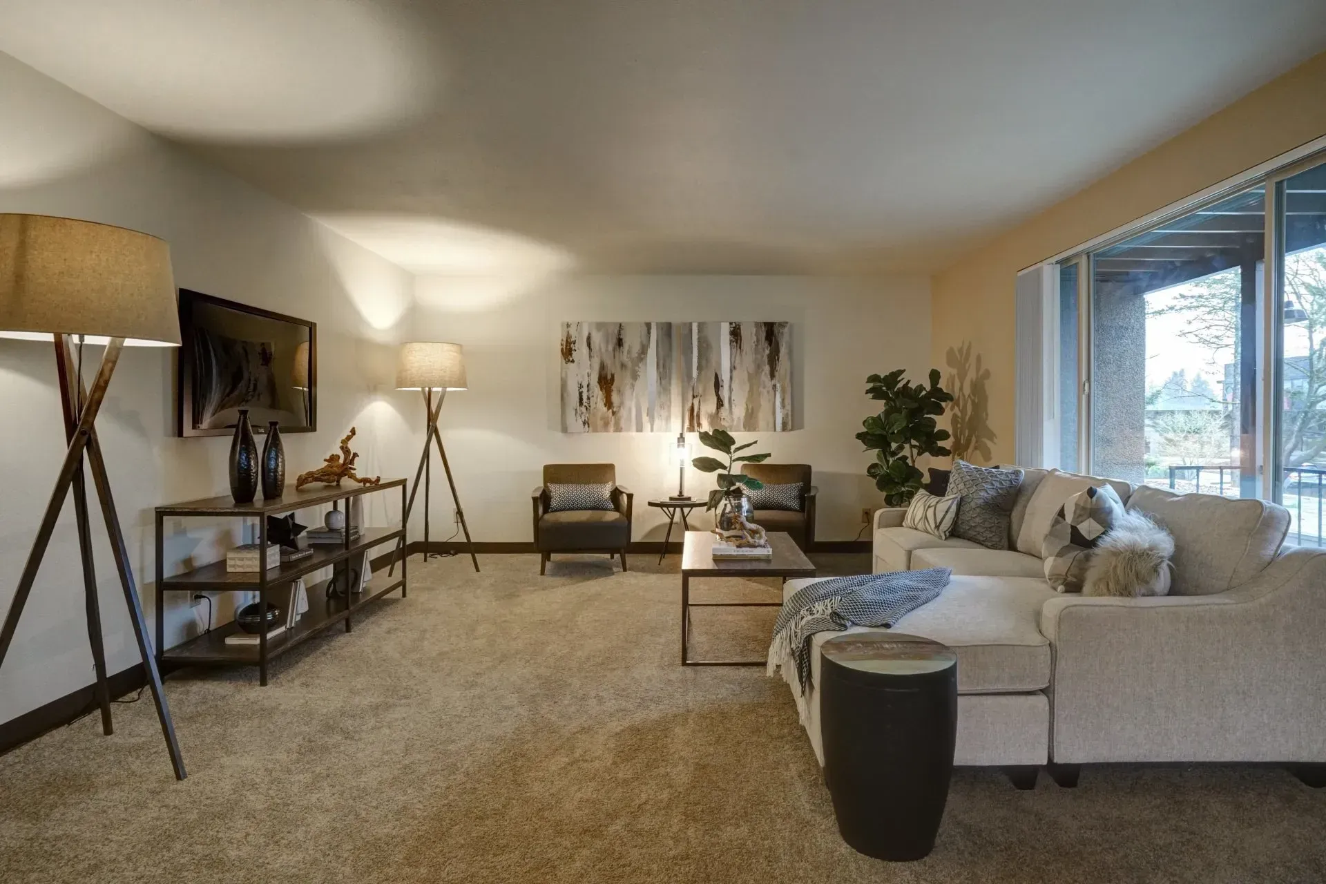 Cozy living room with beige carpet, sectional sofa, artwork, lamps, and a view through a large window.