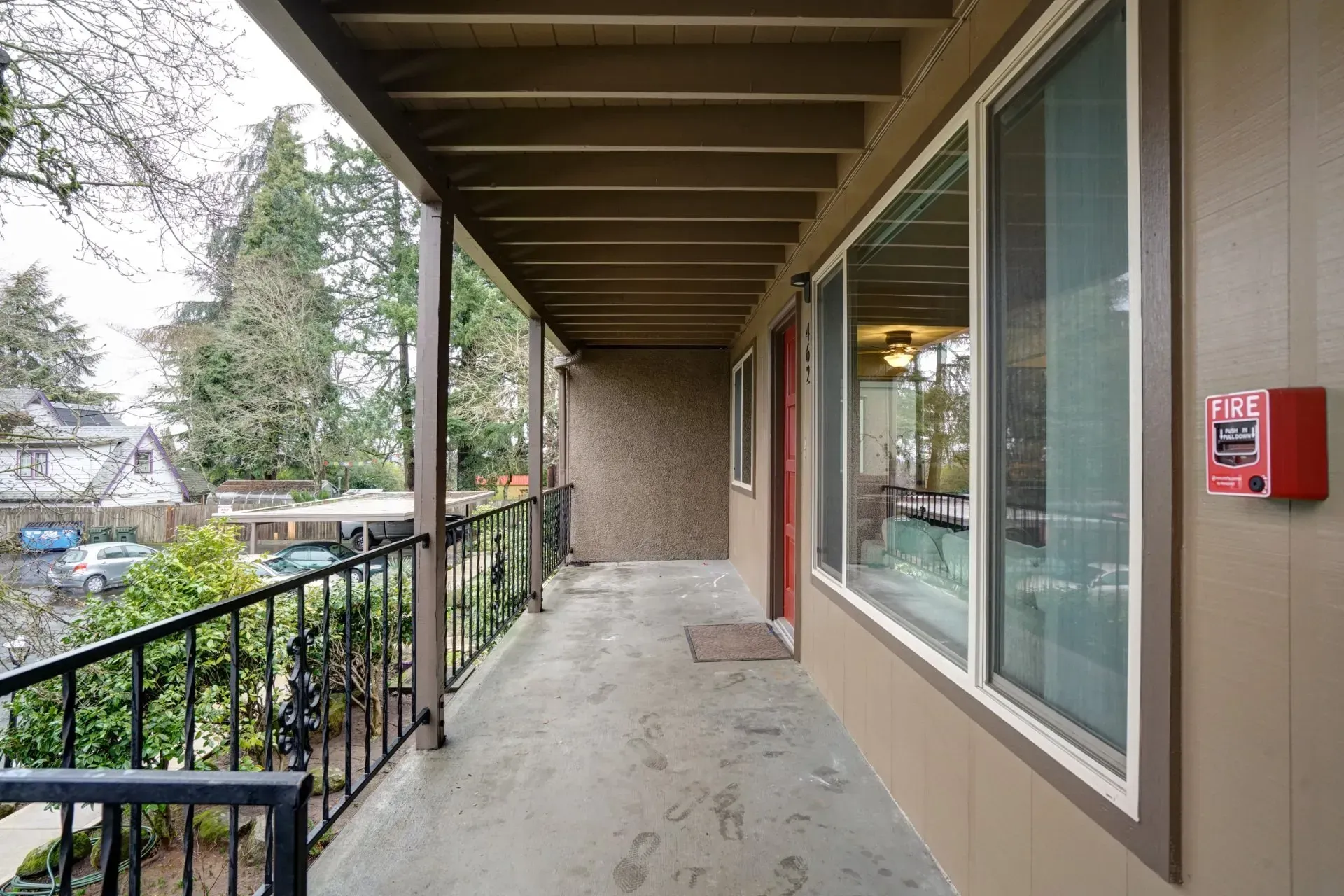 Covered apartment balcony with black railing, large windows, and a red fire alarm.