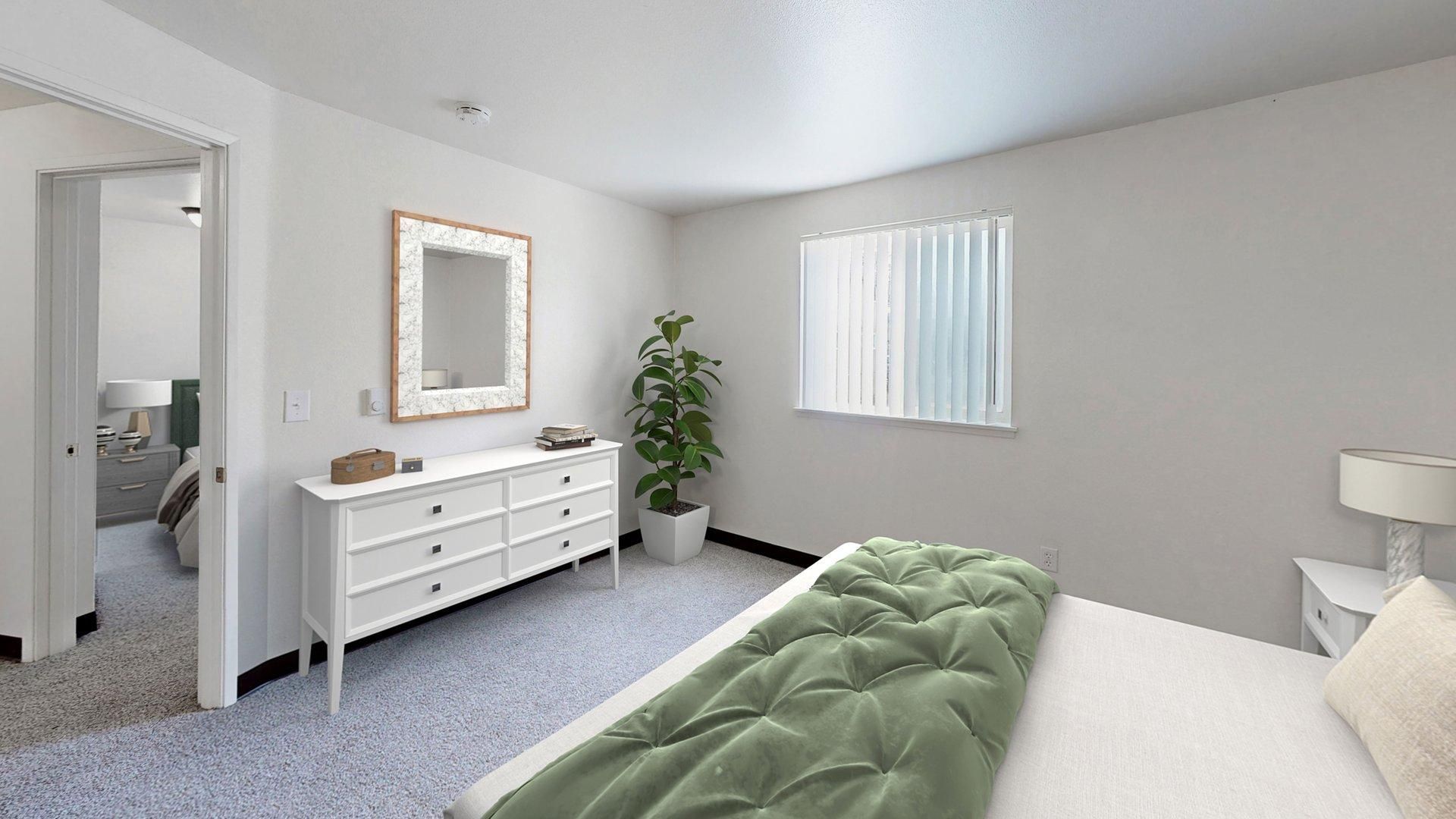 Bedroom with white dresser, mirror, bed, and plant. Gray carpet and walls; open doorway to another room.