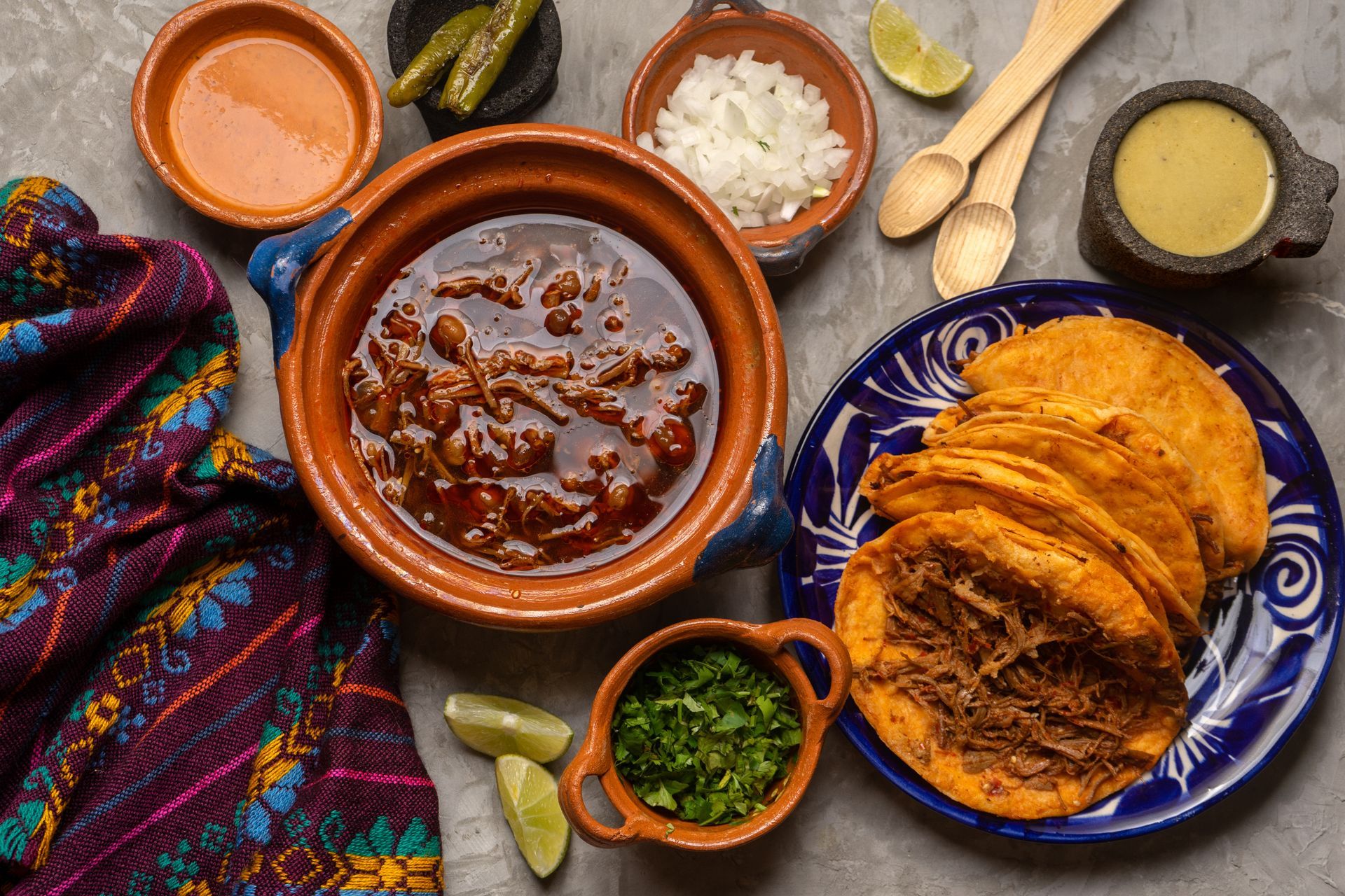 Mexican birria tacos with consommé, condiments, and tortillas on a table.