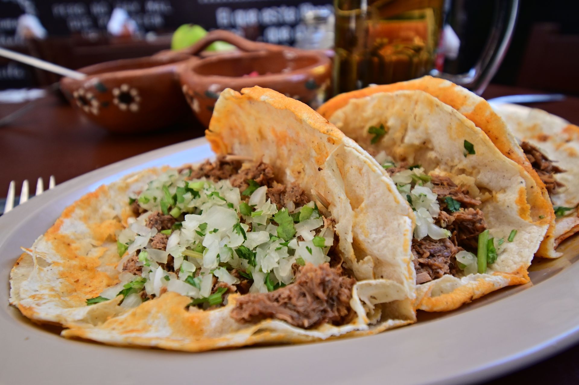 Three tacos with meat, onions, and cilantro on a plate, with a beer and salsa in the background.