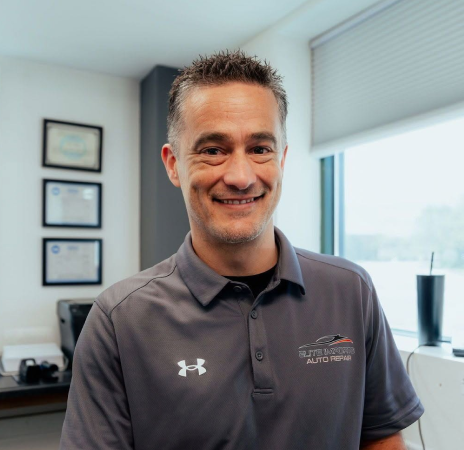 Smiling man in a gray polo shirt standing in a bright office near framed certificates and a window