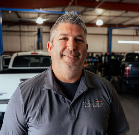 Smiling man in a gray polo shirt standing in a garage with vehicles in the background