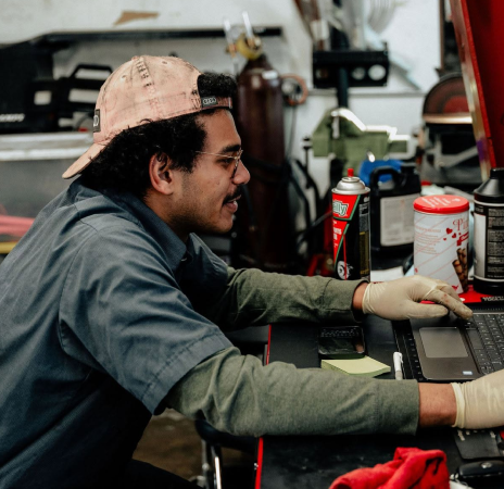 Mechanic in a shop working on a laptop beside tools and spray cans