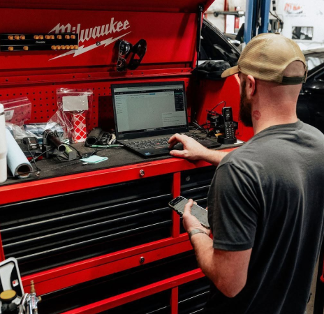 Mechanic in a red Milwaukee tool chest workspace using a laptop, with tools and parts spread across the bench