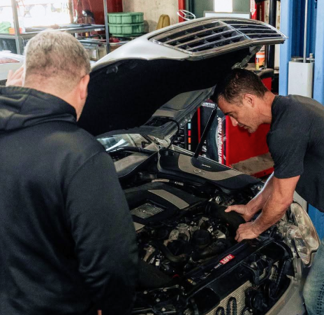 Two mechanics inspect a black car with its hood open in an auto shop.