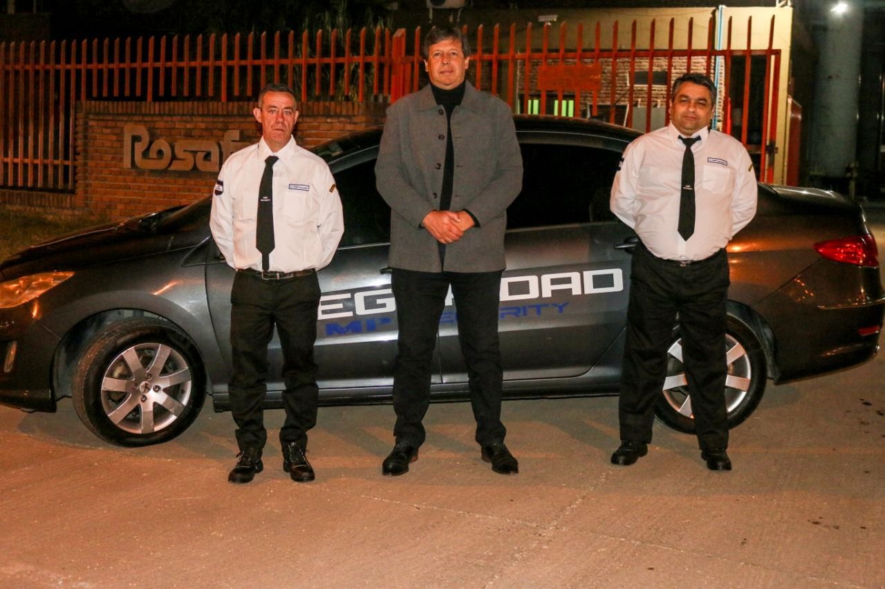Tres hombres uniformados están de pie frente a un coche; dos son guardias. Escenario nocturno, colores oscuros.