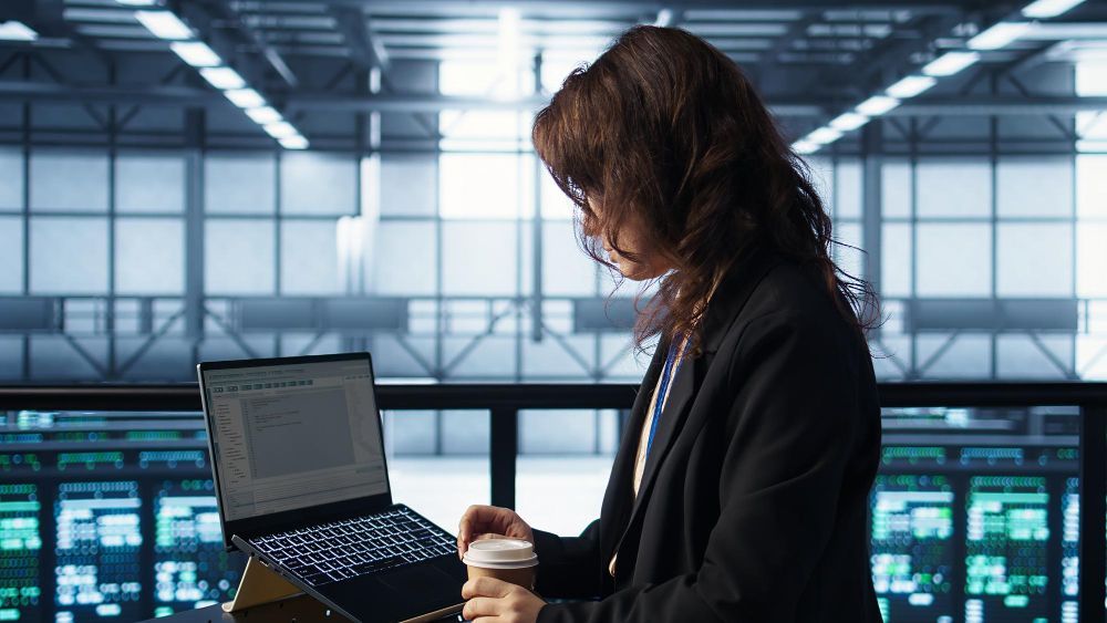 Woman in black blazer working on laptop, holding coffee, in modern office.