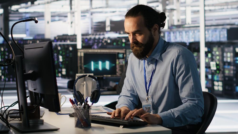 Man with beard typing at computer in server room.
