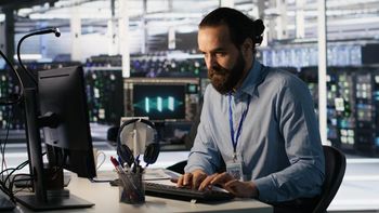Man with beard typing at computer in server room.