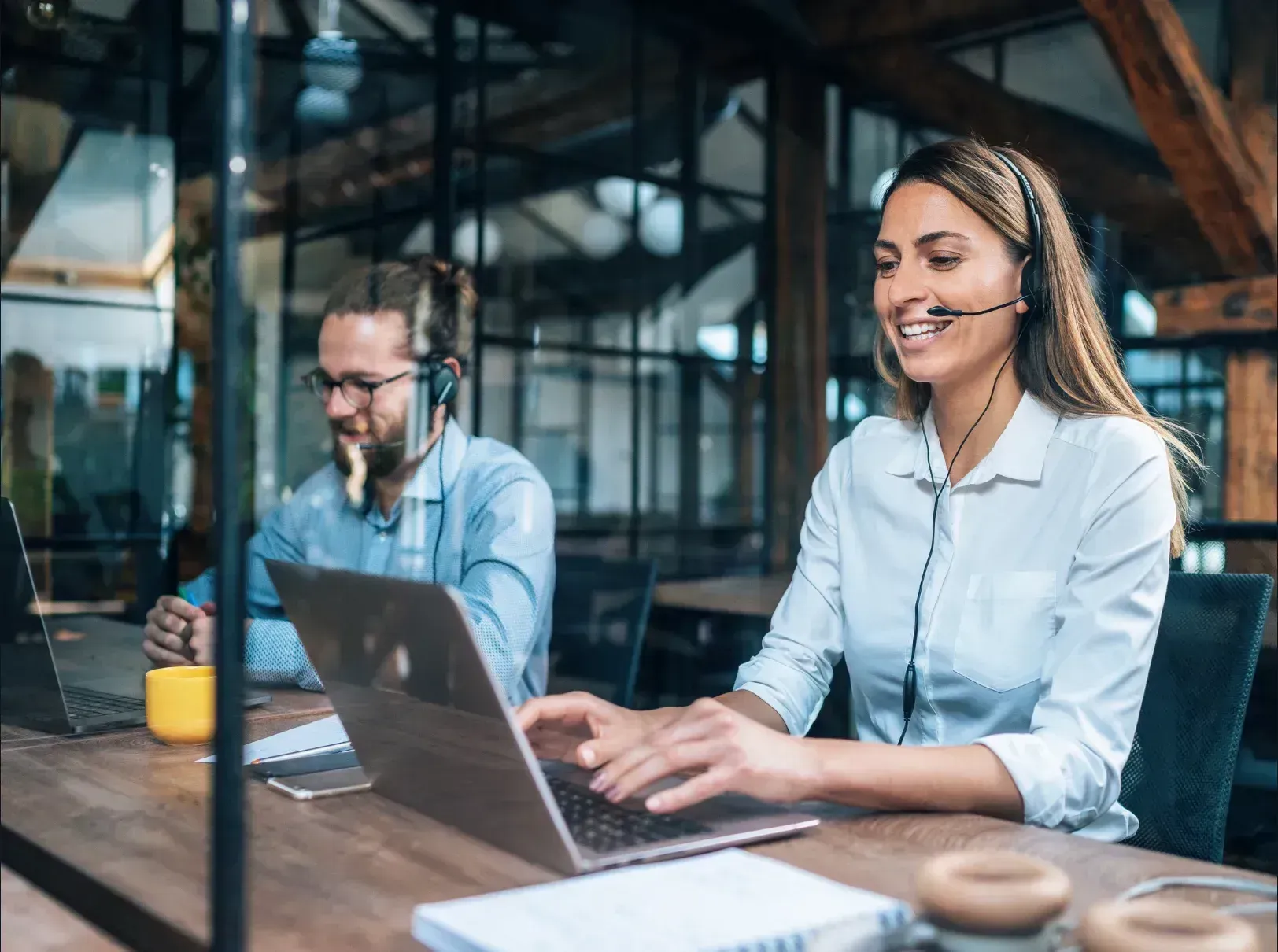 Two people wearing headsets, working on laptops at a wooden table in an office setting.
