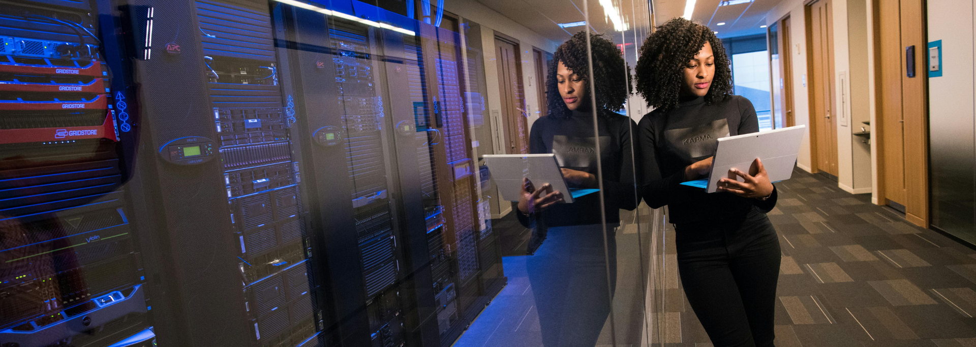 Woman with tablet leaning against glass in a hallway next to server racks.