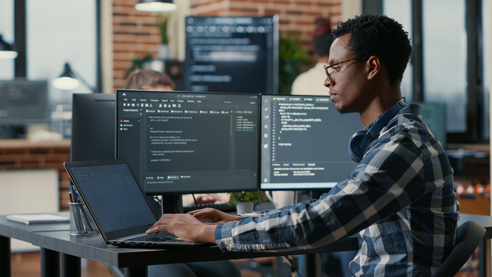 Man in plaid shirt coding on a laptop with two monitors in a modern office.