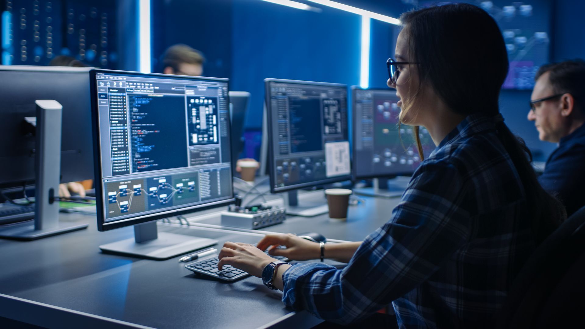 Woman in glasses types at a computer in a server room, smiling, with multiple monitors displaying code and graphics.