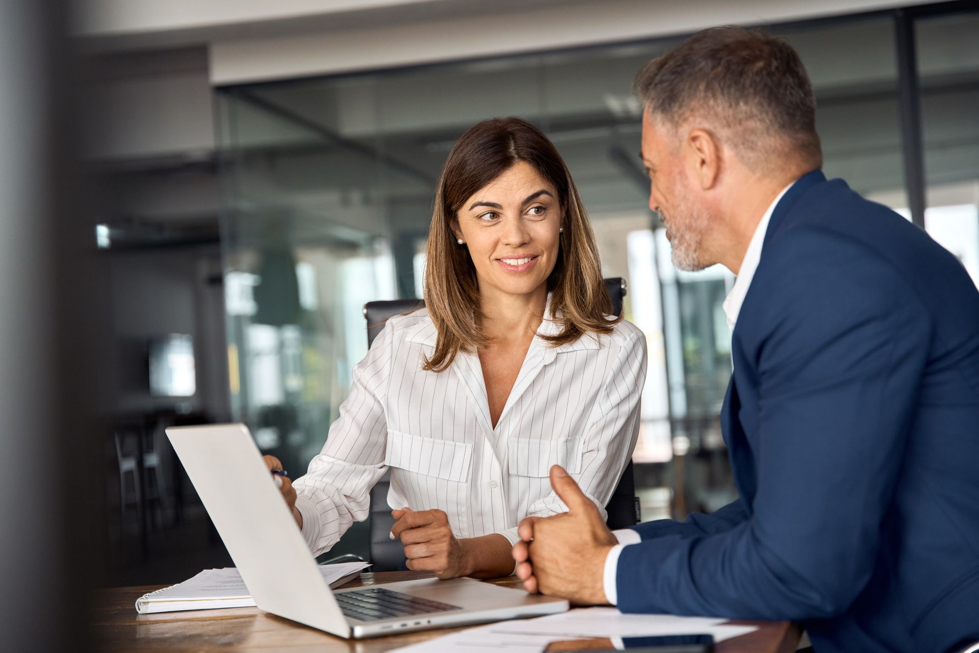 Woman and man in a meeting, woman points at laptop screen, man listens, both at a desk, office setting.