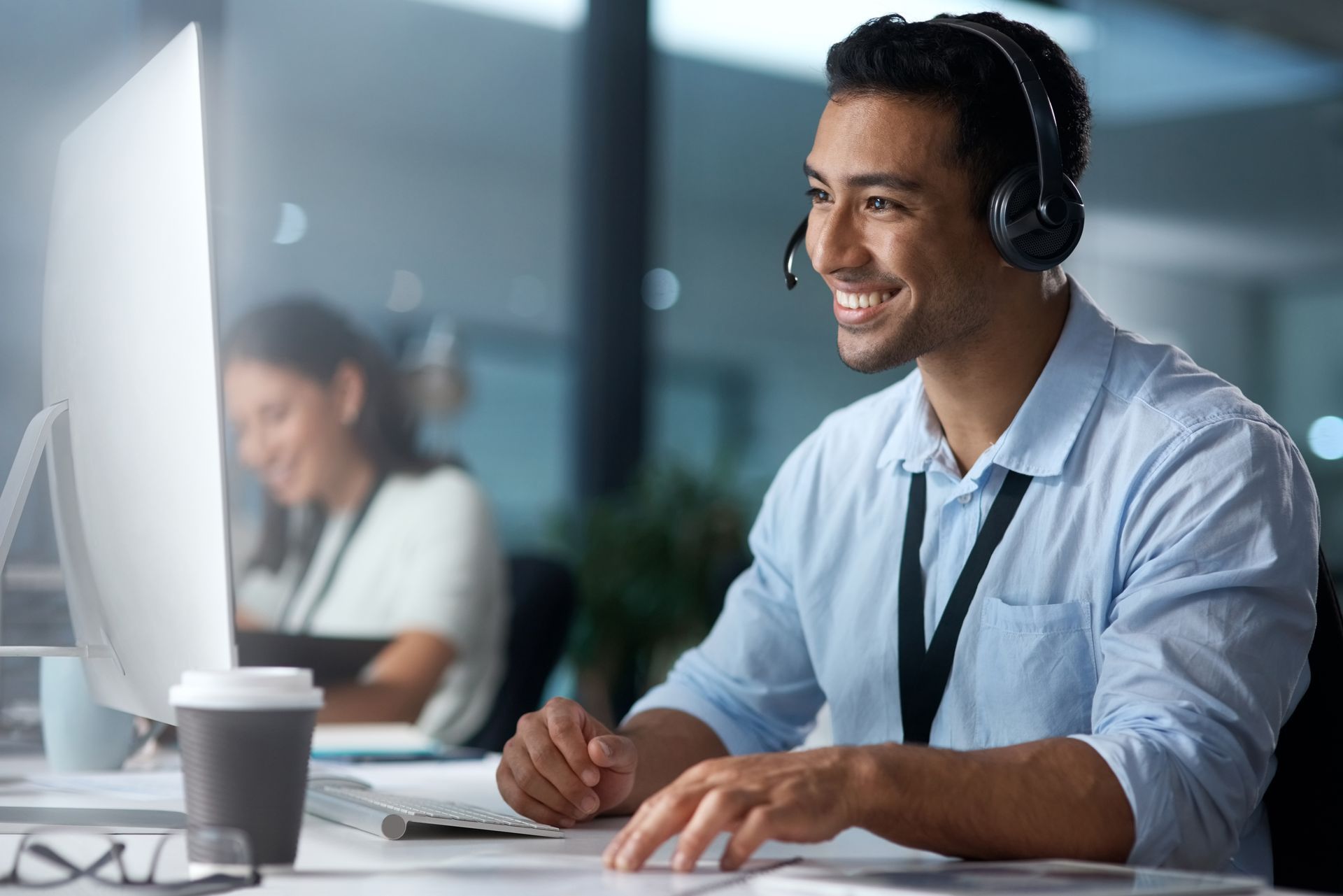 Man with headset smiling, working at computer in office; person in background.