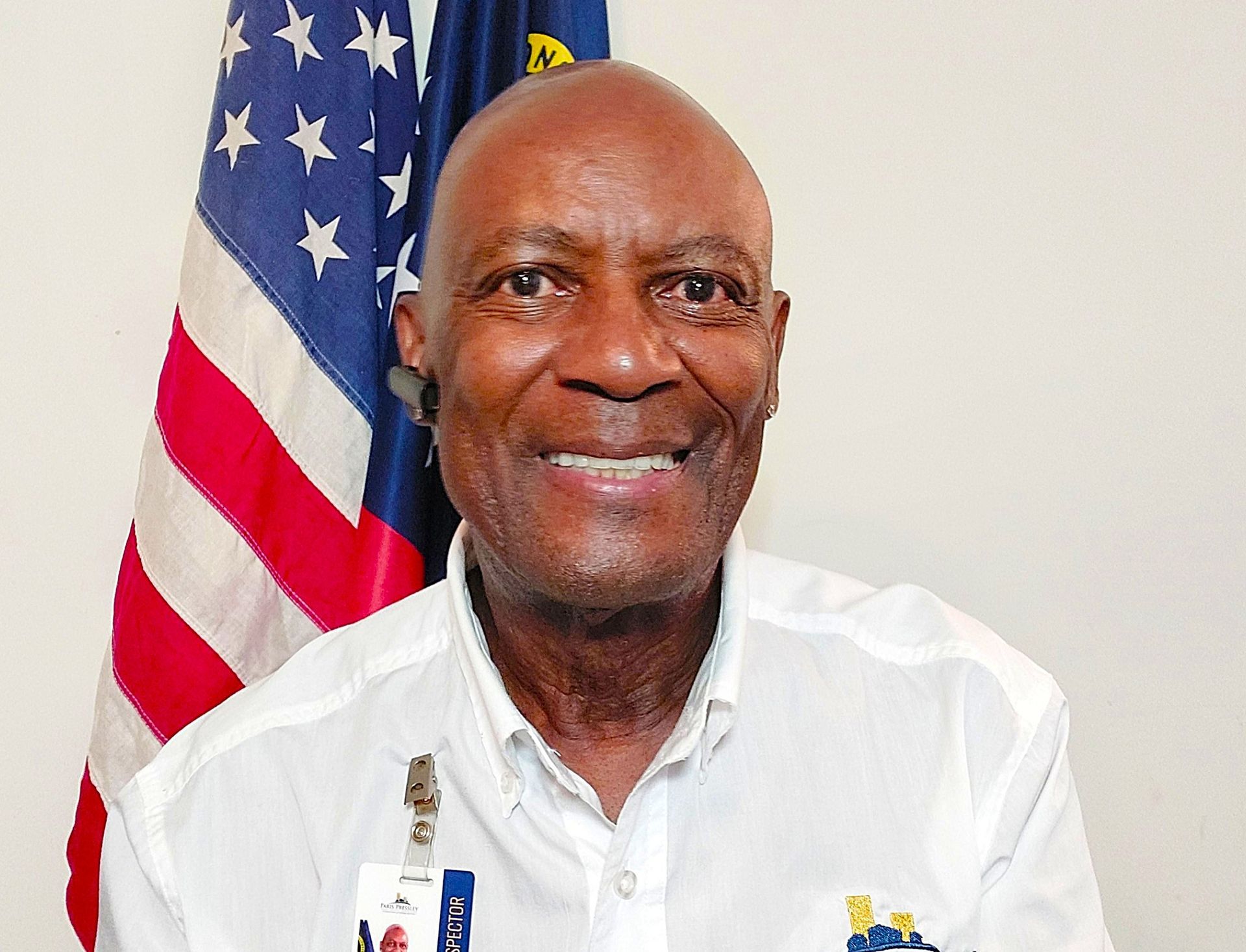 Man smiling, wearing a white shirt, in front of an American flag.