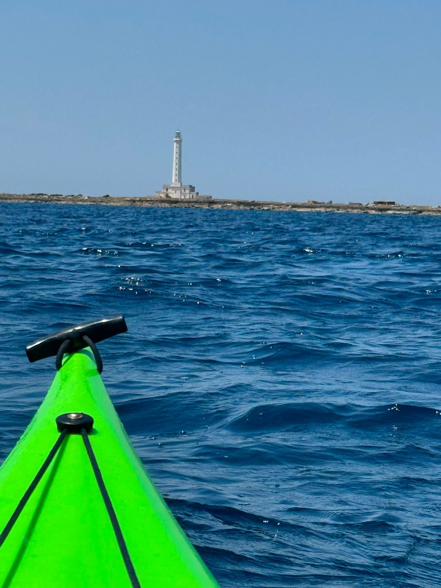 Un kayak verde su un oceano blu si avvicina a un faro lontano sotto un cielo limpido.