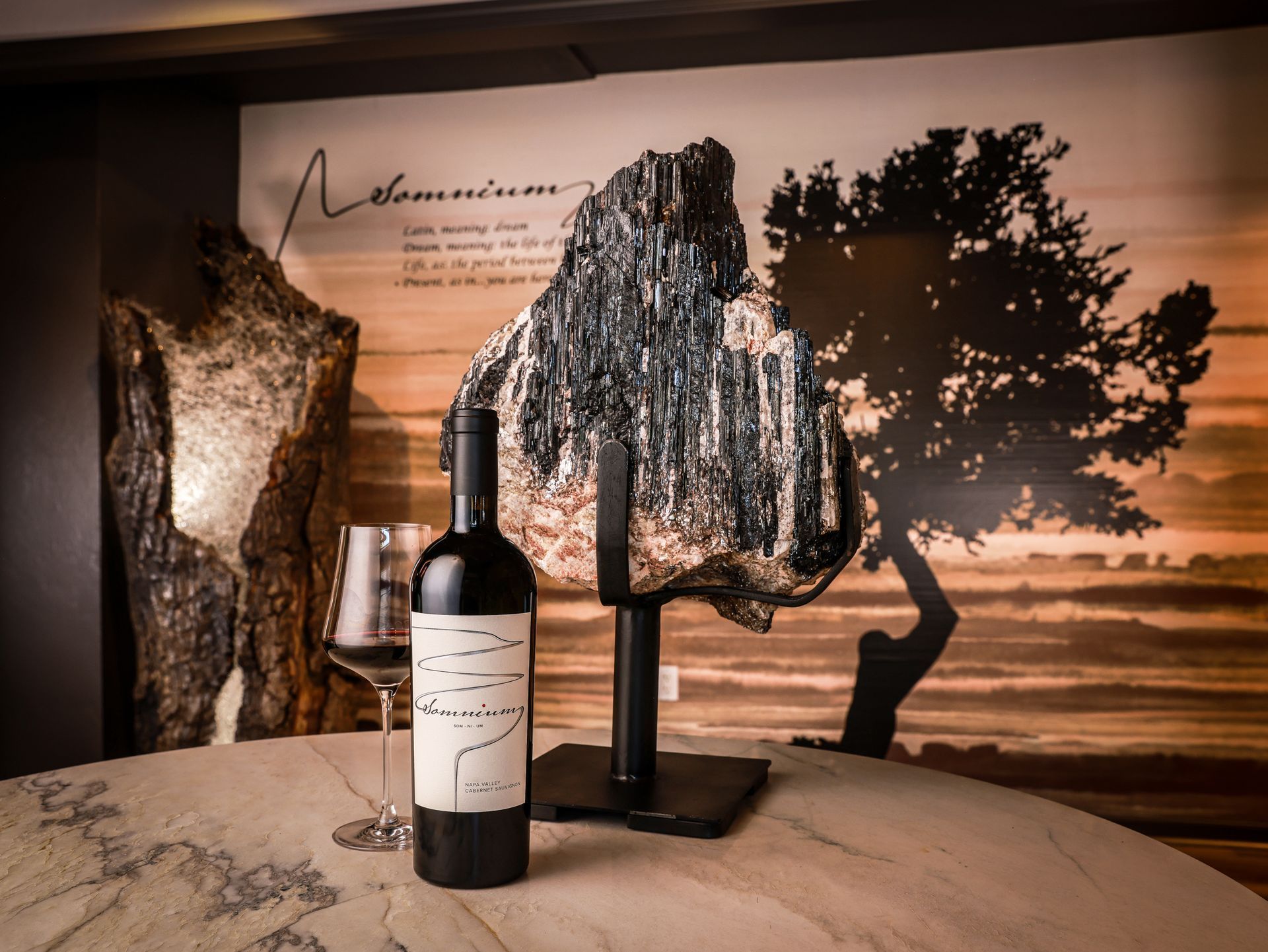 Wine bottle and glass on a table with a rock sculpture in front of a landscape mural.