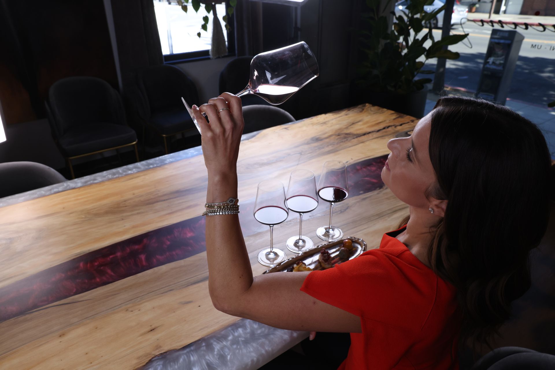 Woman in orange shirt swirling wine, seated at a wooden table with wine glasses, in a cafe setting.