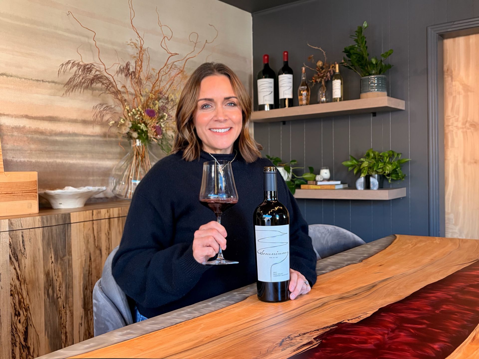 Woman with dark hair smiles, wearing a black sequined top, posing in front of a textured gold wall.