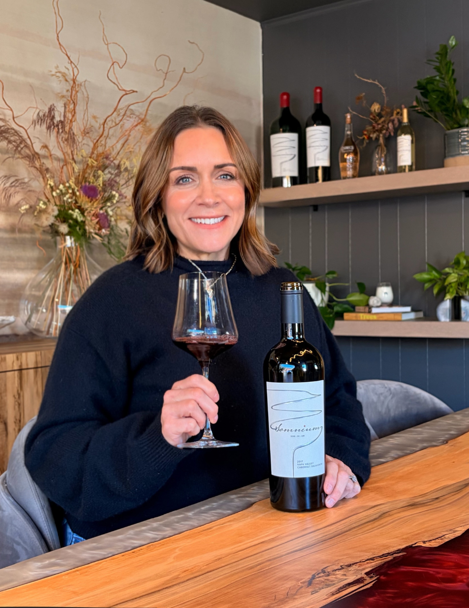 Woman with dark hair smiles, wearing a black sequined top, against a mottled brown background.