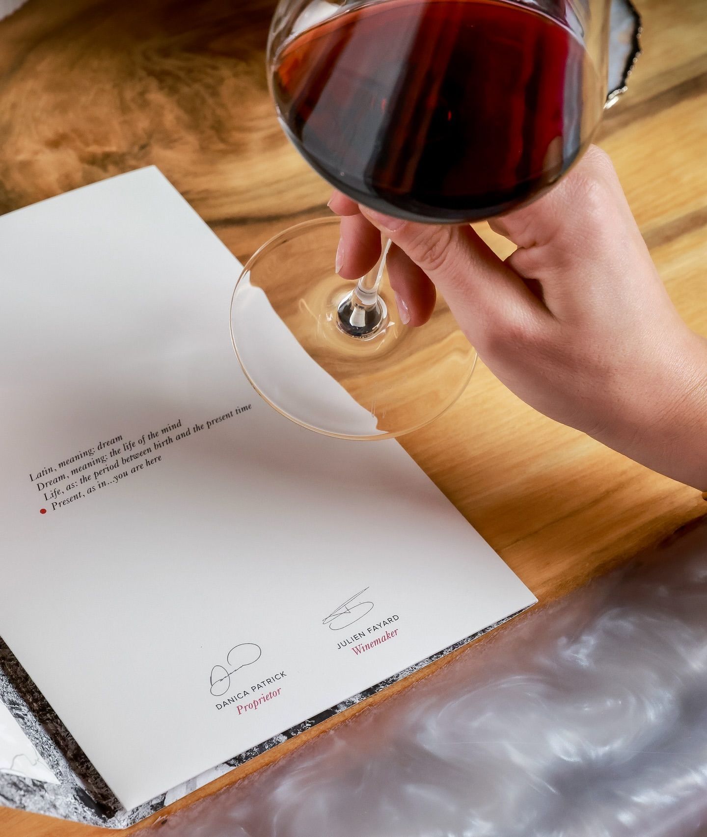 A hand holds a wine glass over a menu on a wooden table. Red wine, text, and signatures are visible.