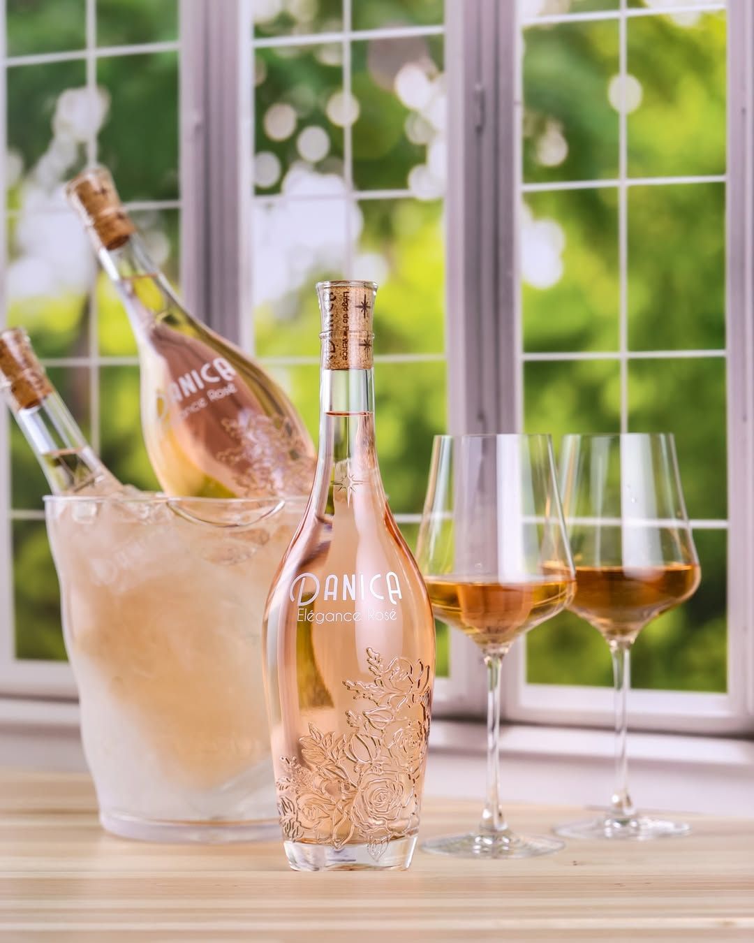 Rose wine bottles in an ice bucket, with two filled glasses and a window background.
