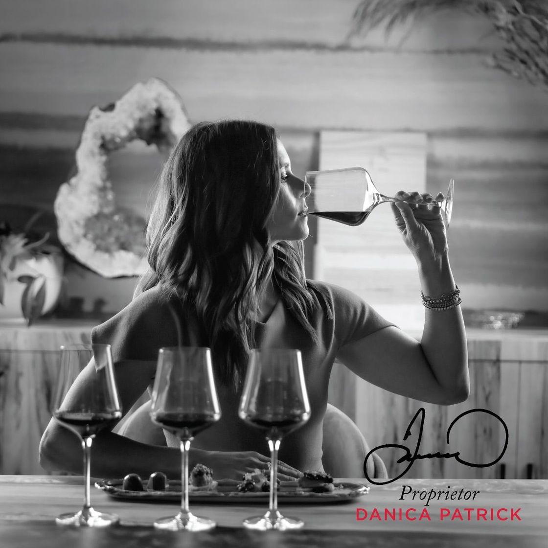 Woman enjoying wine, sitting at a table with glasses and food. Signature and 