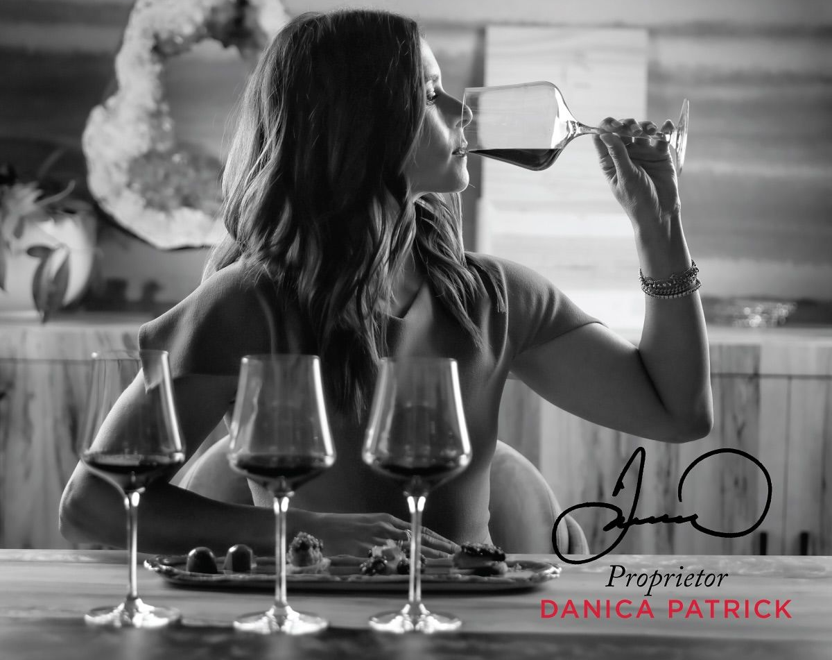 Woman enjoying wine, sitting at a table with glasses and food. Signature and 