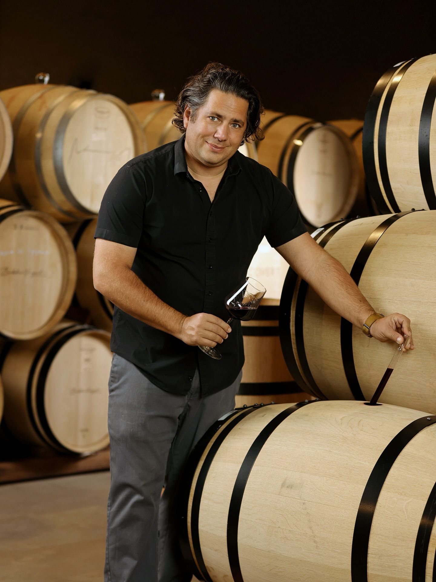 Man in black shirt holding a wine glass, standing in a cellar with wine barrels.